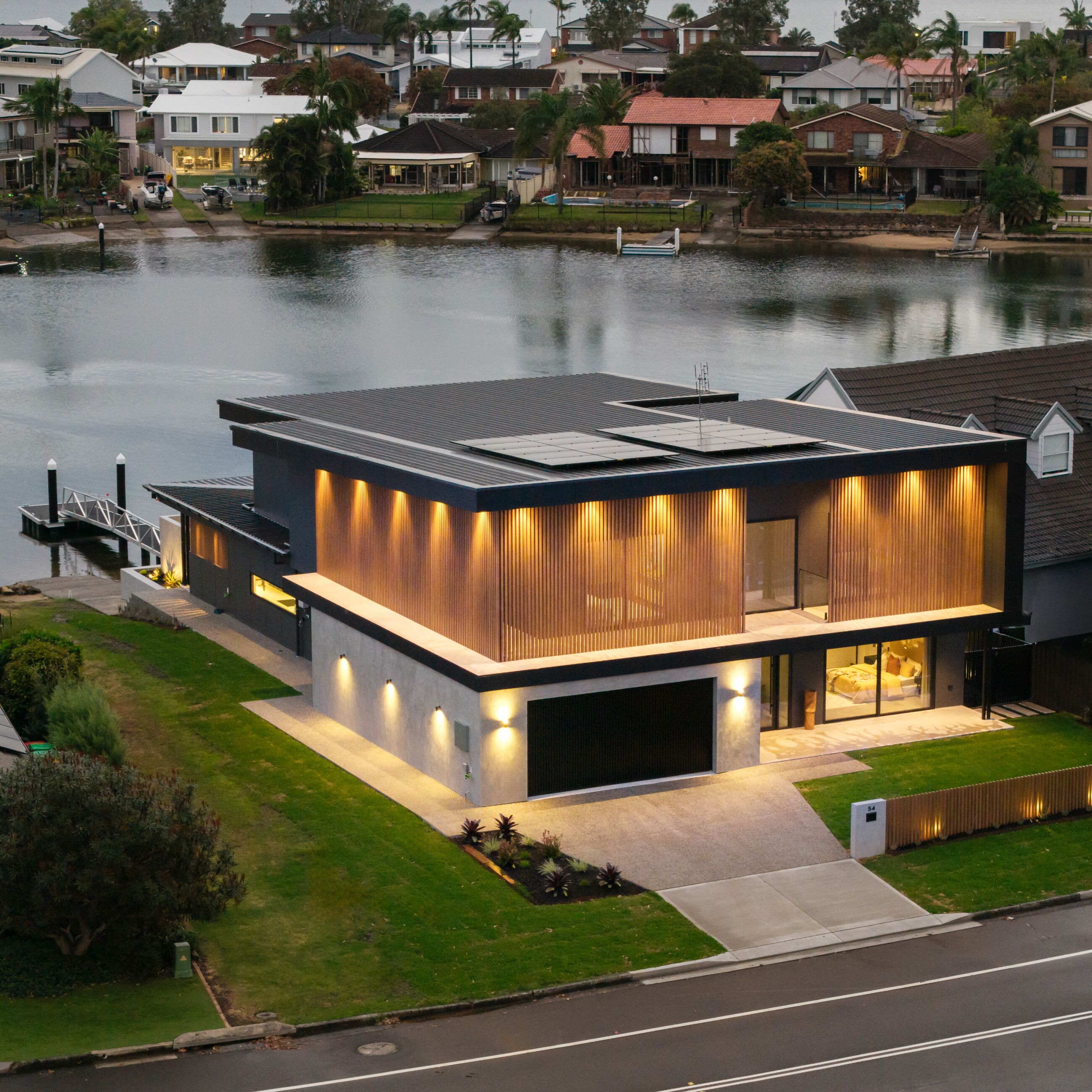 Aerial view of a waterfront home at dusk with facade, garden and interior lighting on